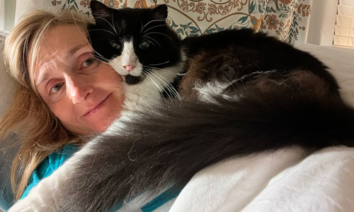 A woman with blonde hair, lying with her head on a pillow. There is a long-haired black and white cat sitting on her chest and shoulder with his head pressed against her face.