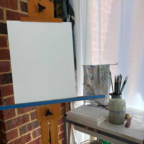 A easel with a blank canvas beside a cart with paint brushes, a palette, and art supplies on it.